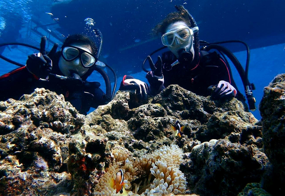 Two people in Scuba gear pose in front of a reef as fish swim in front
